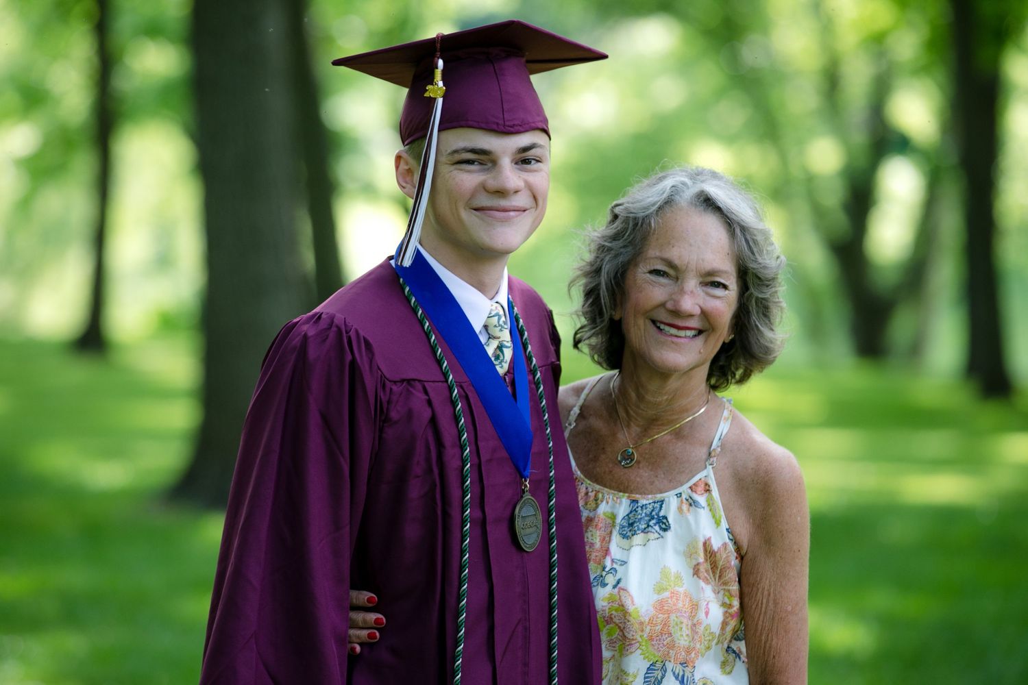 Two graduates in purple caps and gowns pose with others in a sunny outdoor setting with trees in the background.