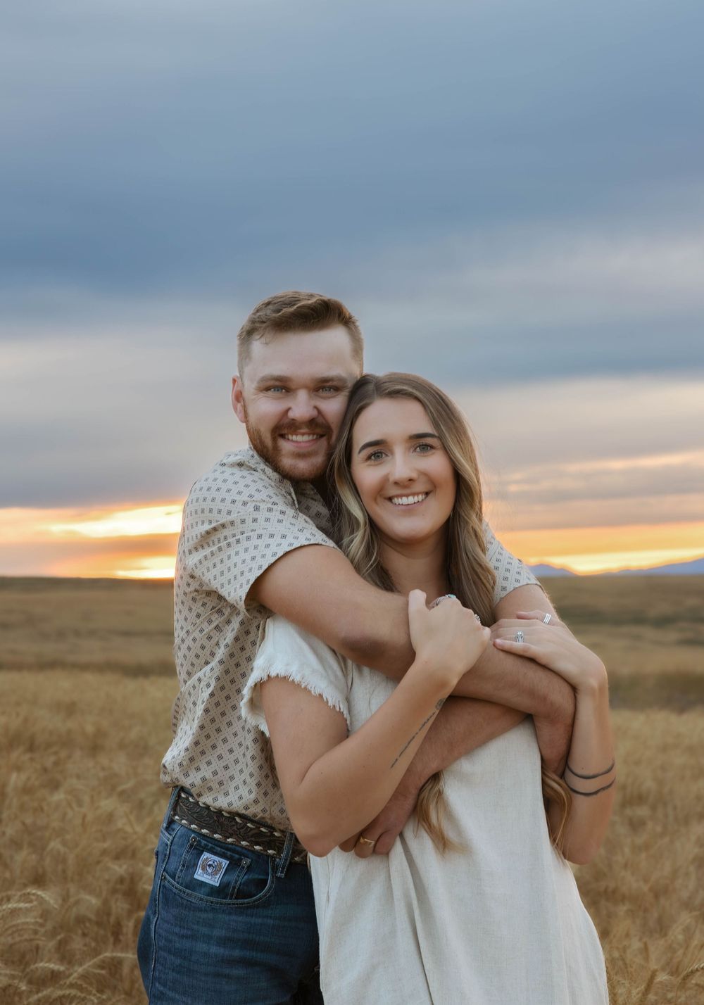 A couple embraces in a wheat field during sunset with dramatic clouds overhead.