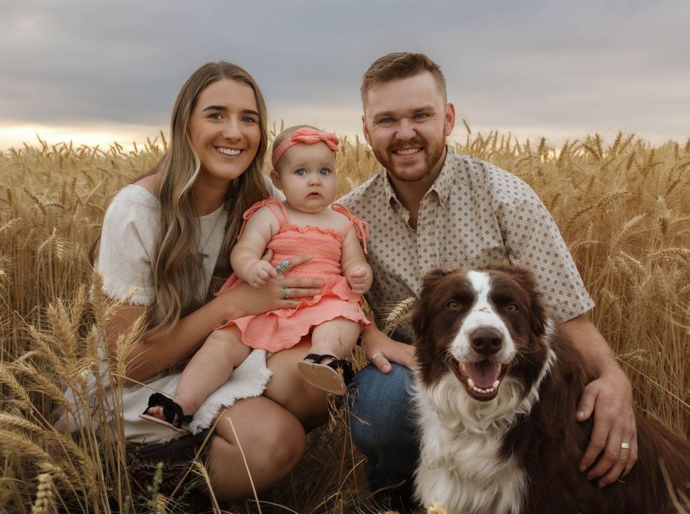 A family and their brown and white dog pose together in a golden wheat field at sunset.