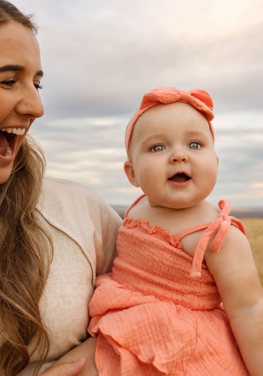 A baby in coral dress smiles while being held in a wheat field at sunset.