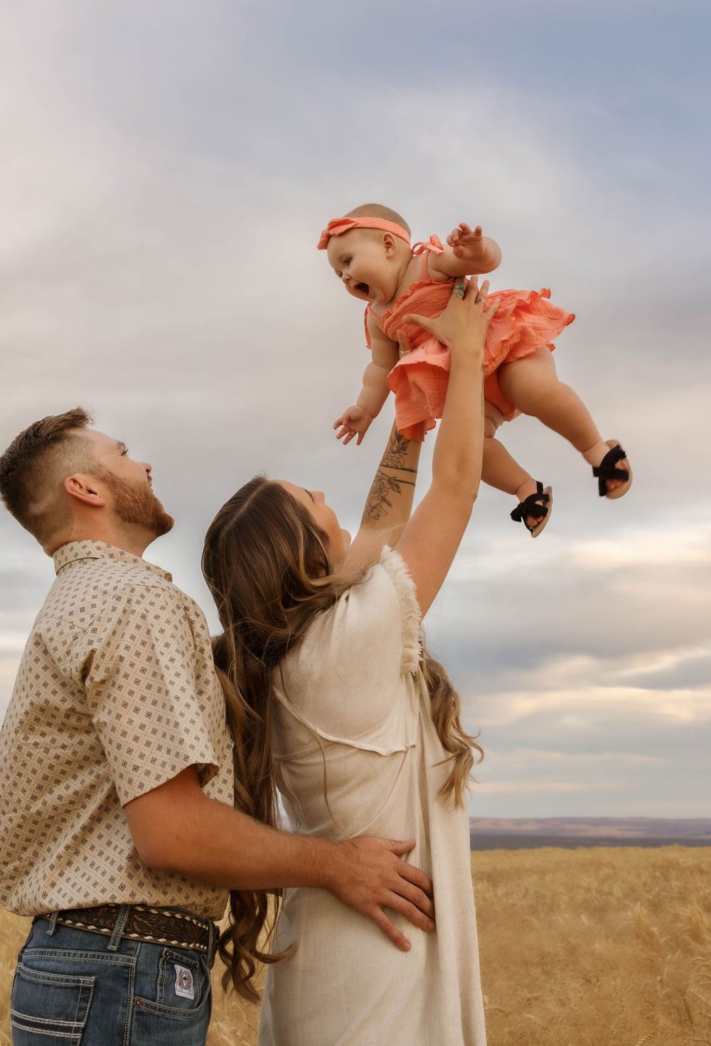 A family shares playful moments lifting their baby in a wheat field at sunset.