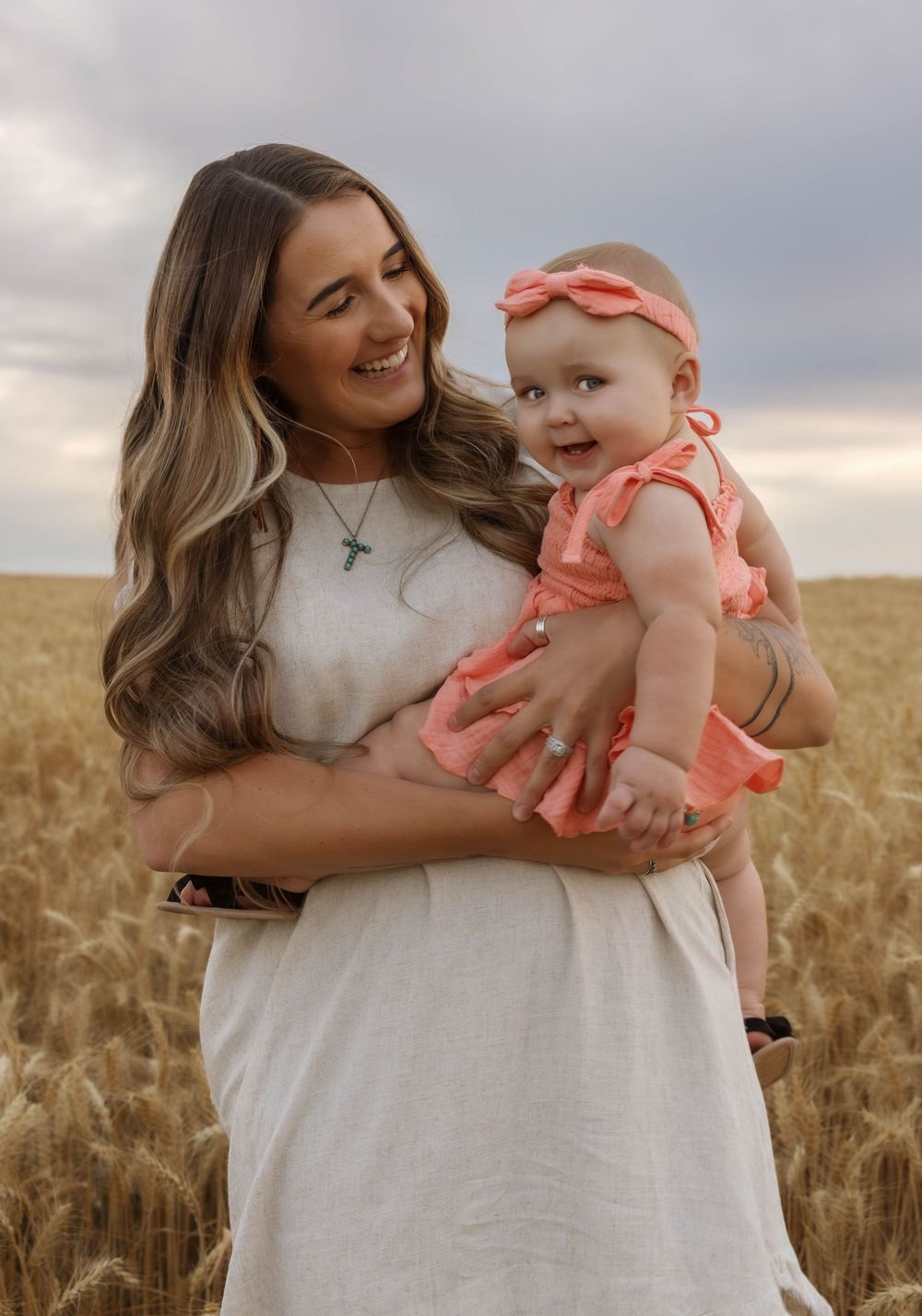A baby wearing coral clothing is held lovingly in a wheat field at sunset.