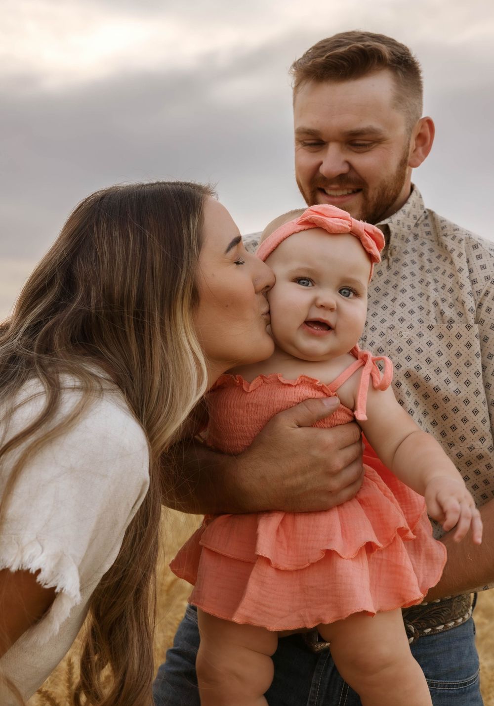 A heartwarming family moment outdoors with a baby in a coral dress being kissed by parents at sunset.
