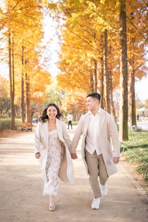 A couple walks hand in hand along a tree-lined path with golden autumn foliage overhead.