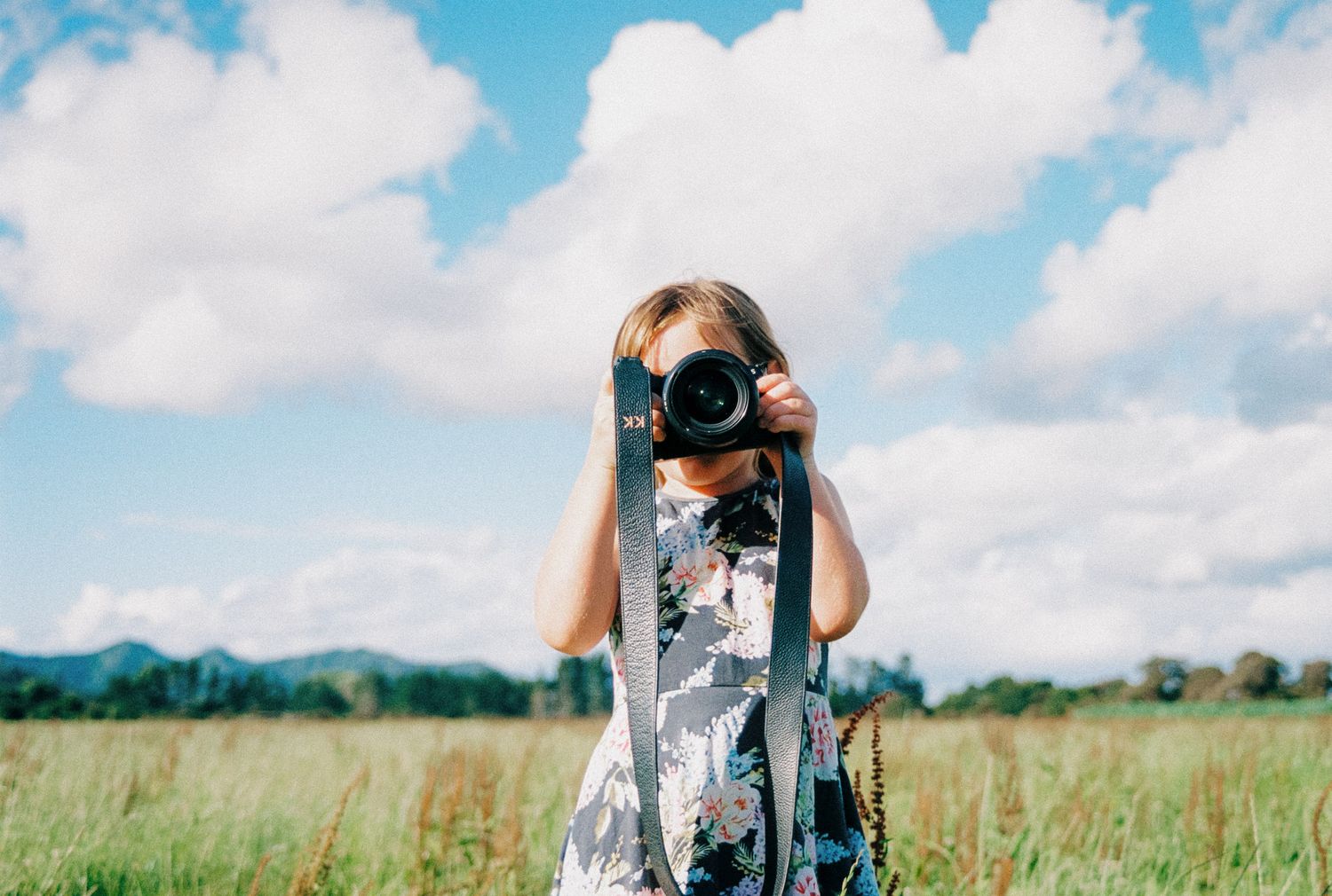 Camera held up to take photos of a scenic mountain landscape under blue sky with puffy white clouds.