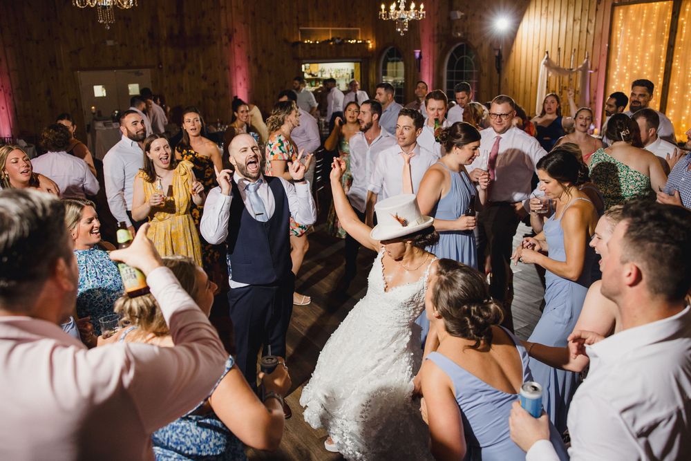 Wedding party guests dancing and celebrating on a crowded rustic barn dance floor beneath warm overhead lighting. RI Wedding Photographer