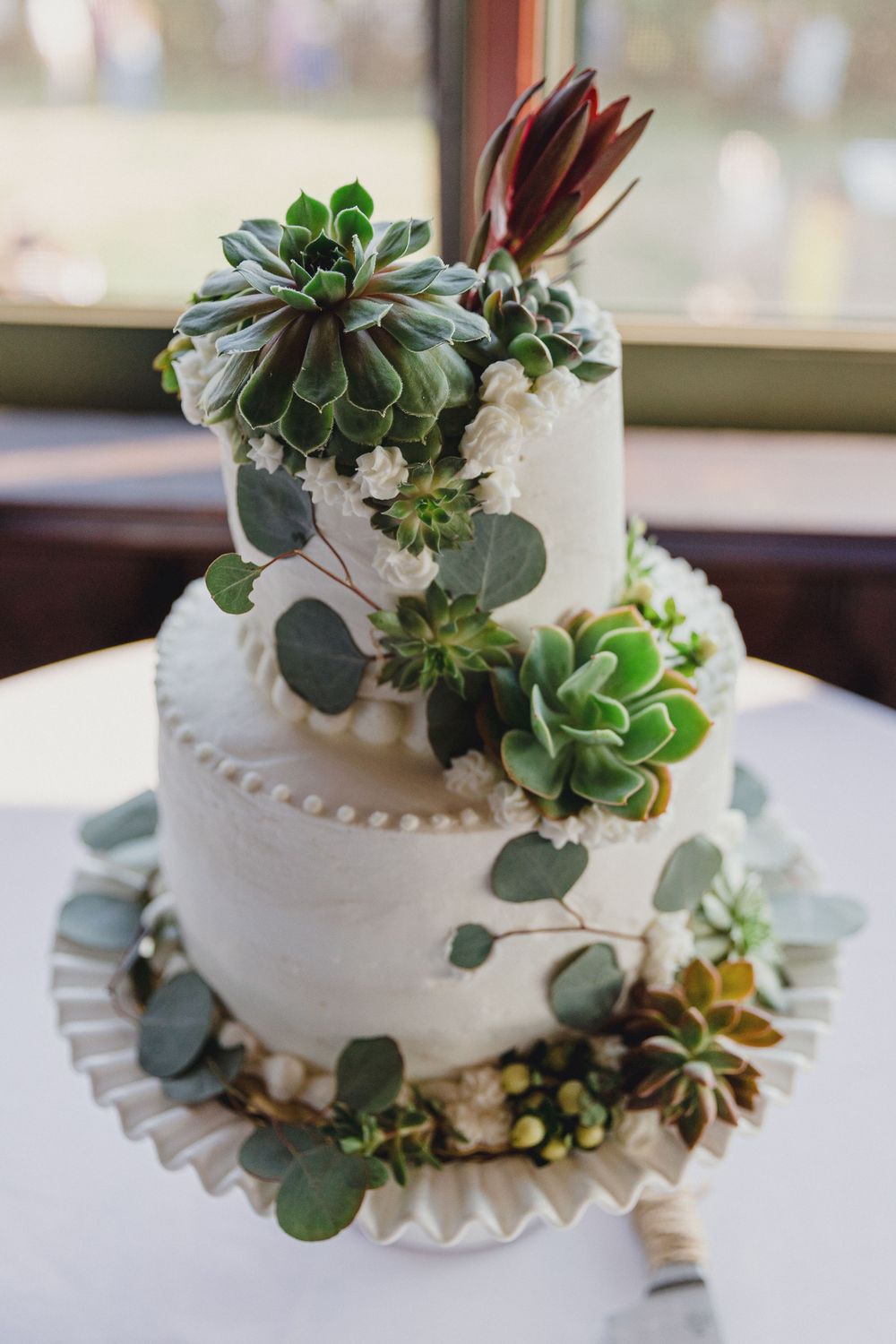 White tiered wedding cake decorated with fresh green succulents and eucalyptus leaves on display by a window. RI Wedding Photographer