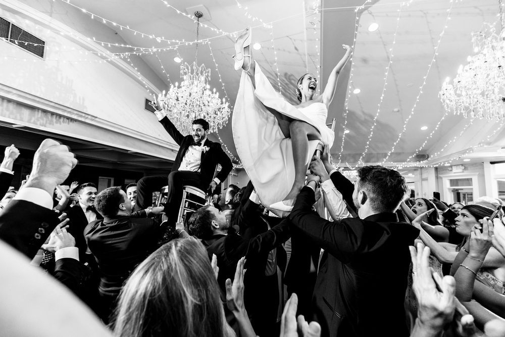 Celebration at a wedding reception as guests lift someone high in the air during lively dance party under chandeliers. RI Wedding Photograph