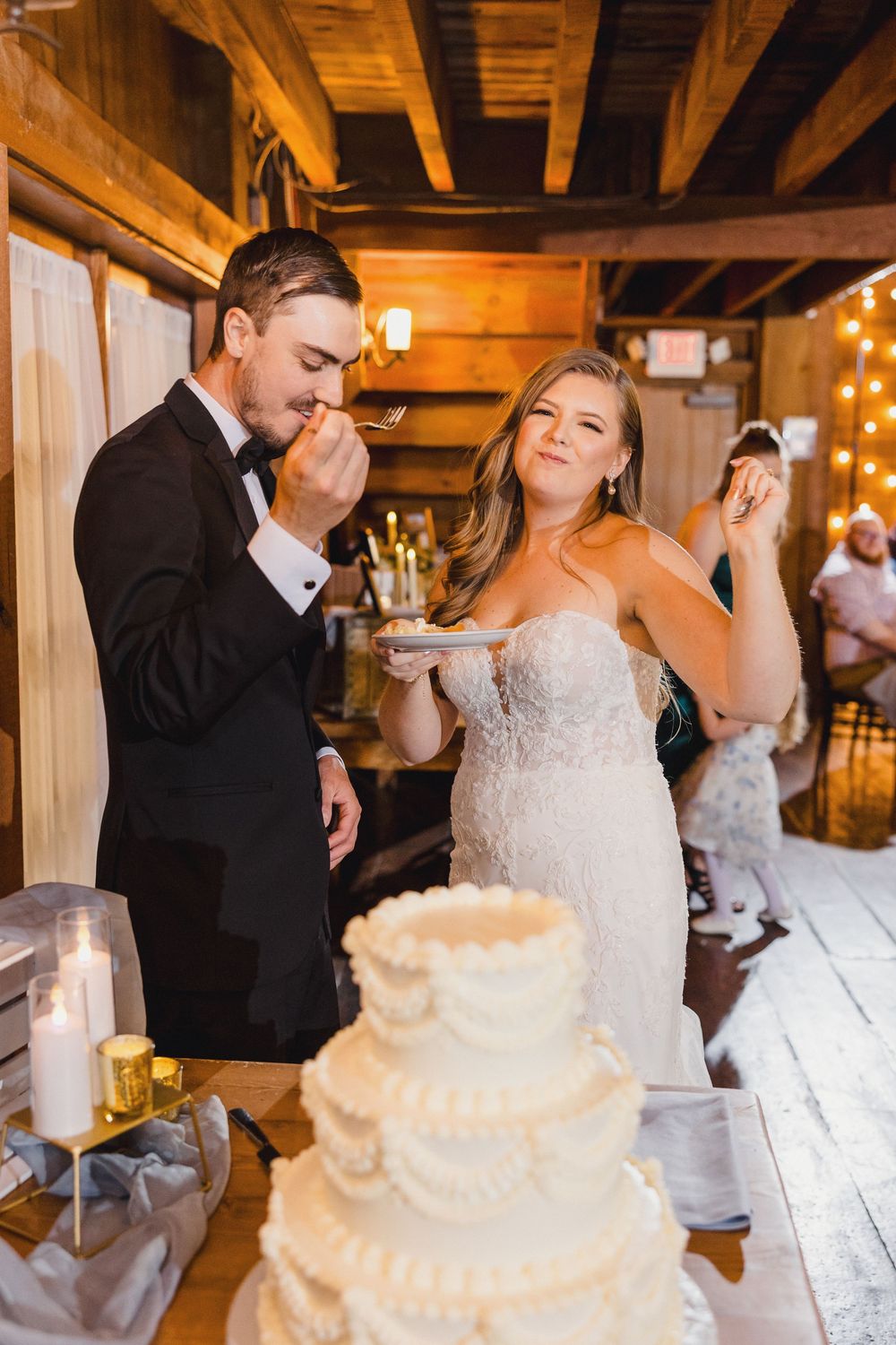 Newlyweds share bites of their elegant white tiered wedding cake during a rustic barn reception celebration. CT wedding Photographer