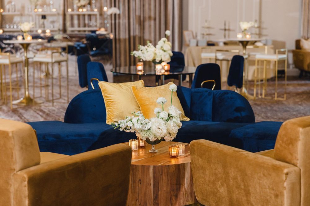 Elegant wedding reception venue with navy blue velvet chairs and gold accents around circular tables with floral centerpieces. Delmar hotel