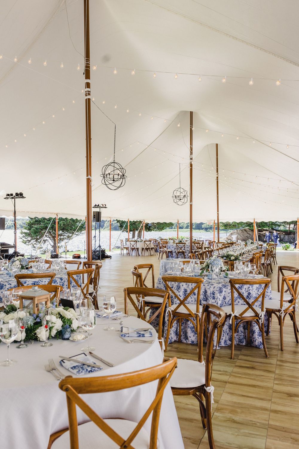 Elegant wedding reception setup under a white tent with round tables, wooden cross-back chairs, and string lights overhead. RI Wedding Photo