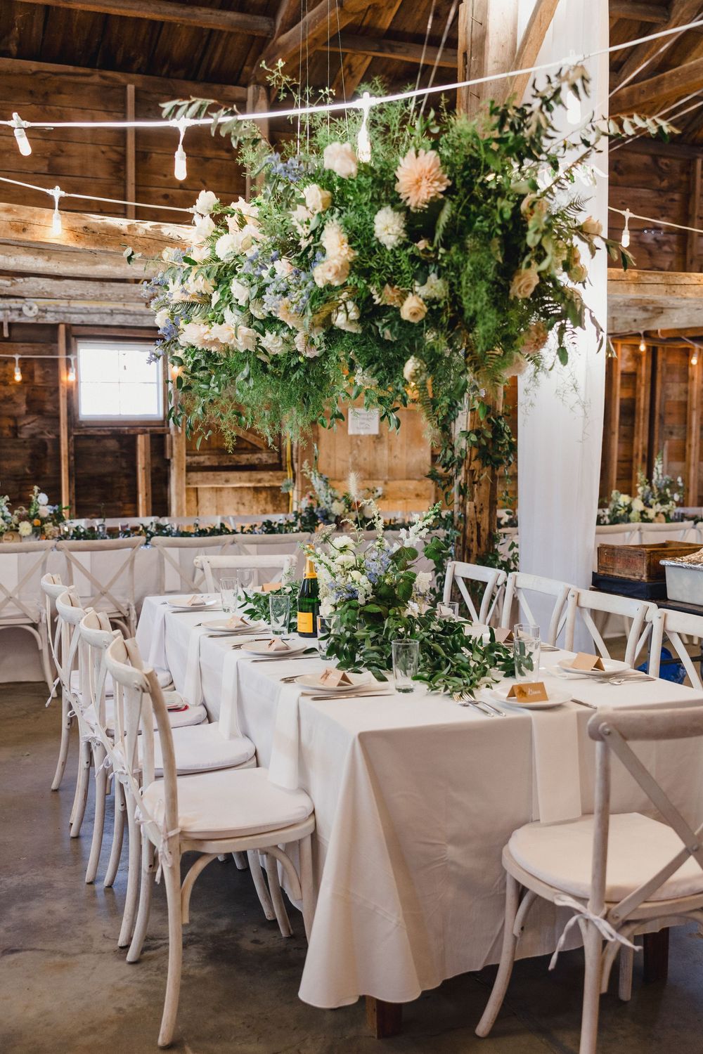 A rustic barn wedding reception setup with elegant white tablecloths, lush floral arrangements overhead. Destination Wedding photographer