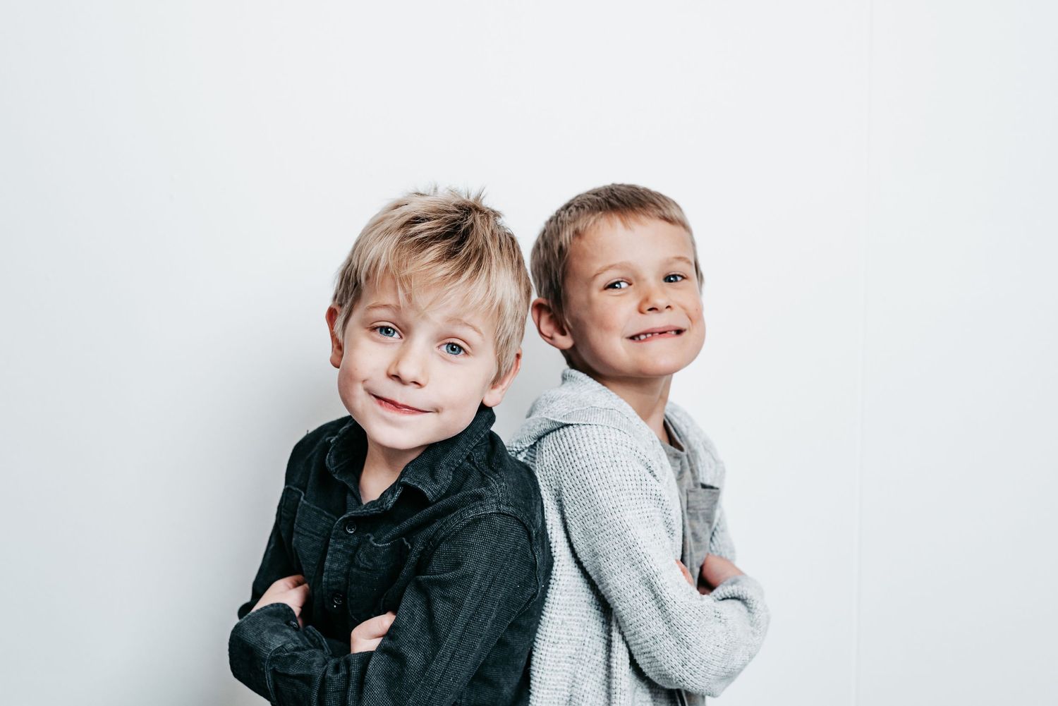 Two children wearing black and gray sweaters lean against each other playfully in a bright white studio setting.