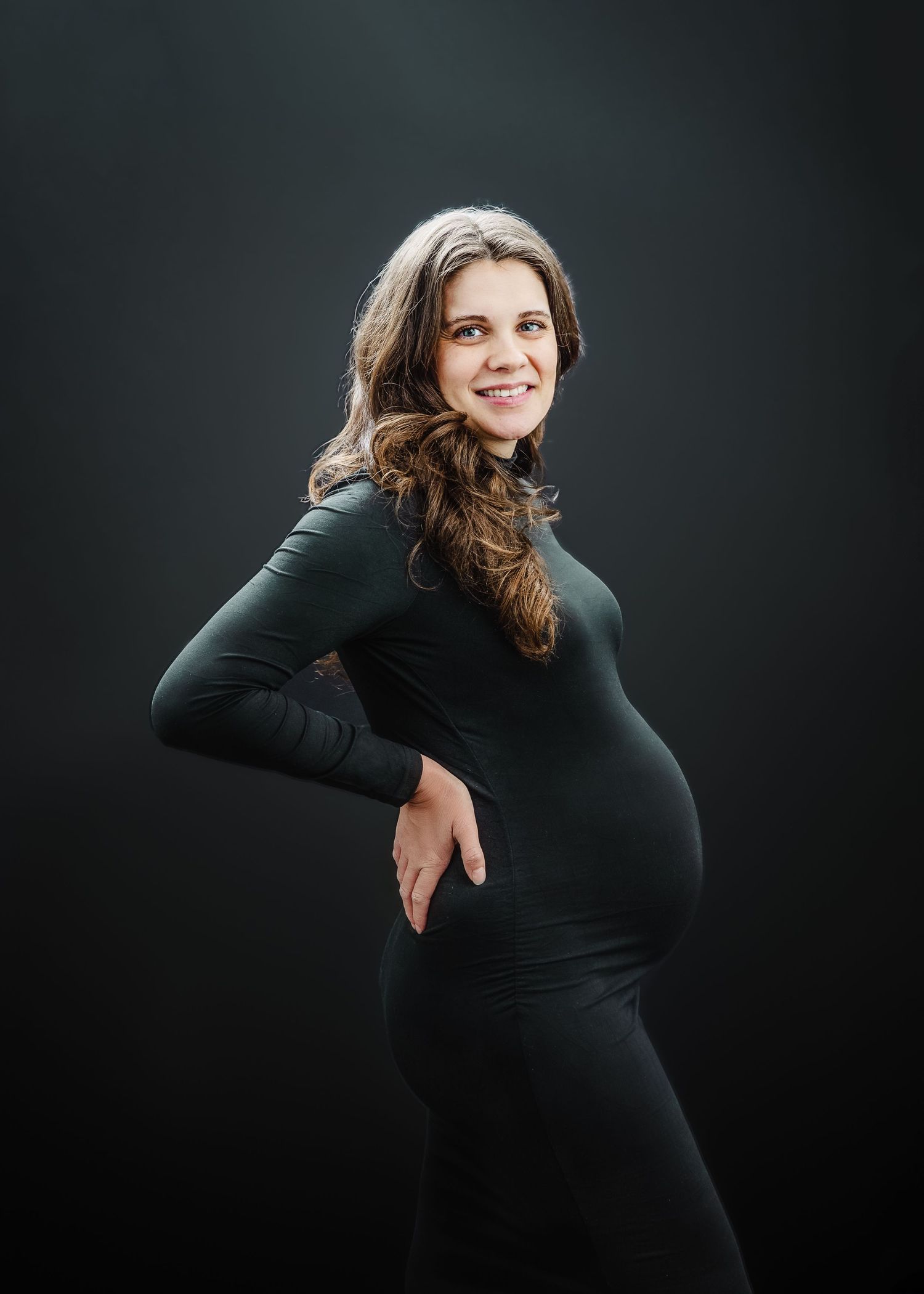 Elegant maternity portrait featuring a long-sleeved black dress against a dark dramatic background.