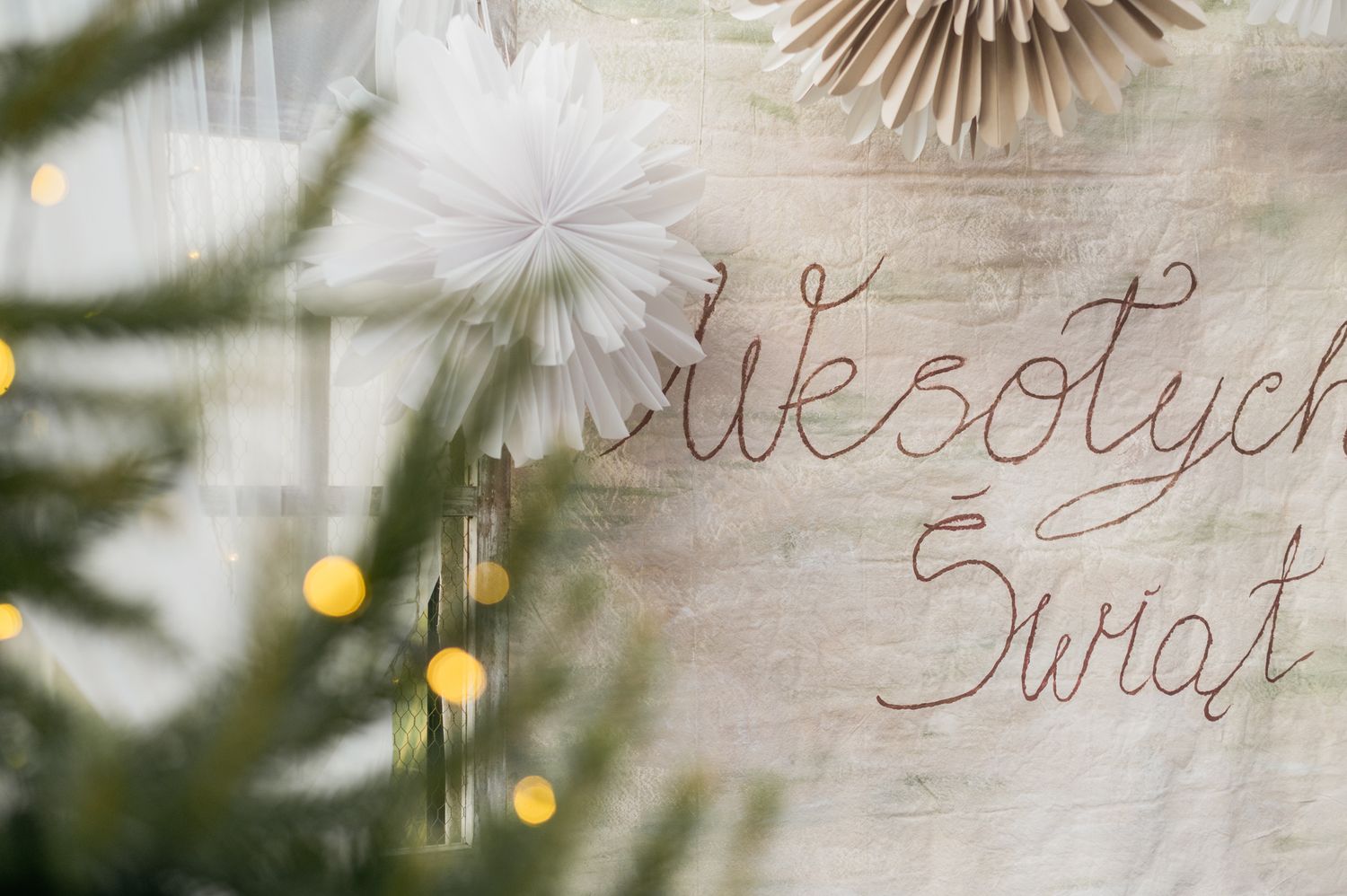 Handwritten calligraphy text on a rustic white wooden background with blurred holiday lights and greenery.