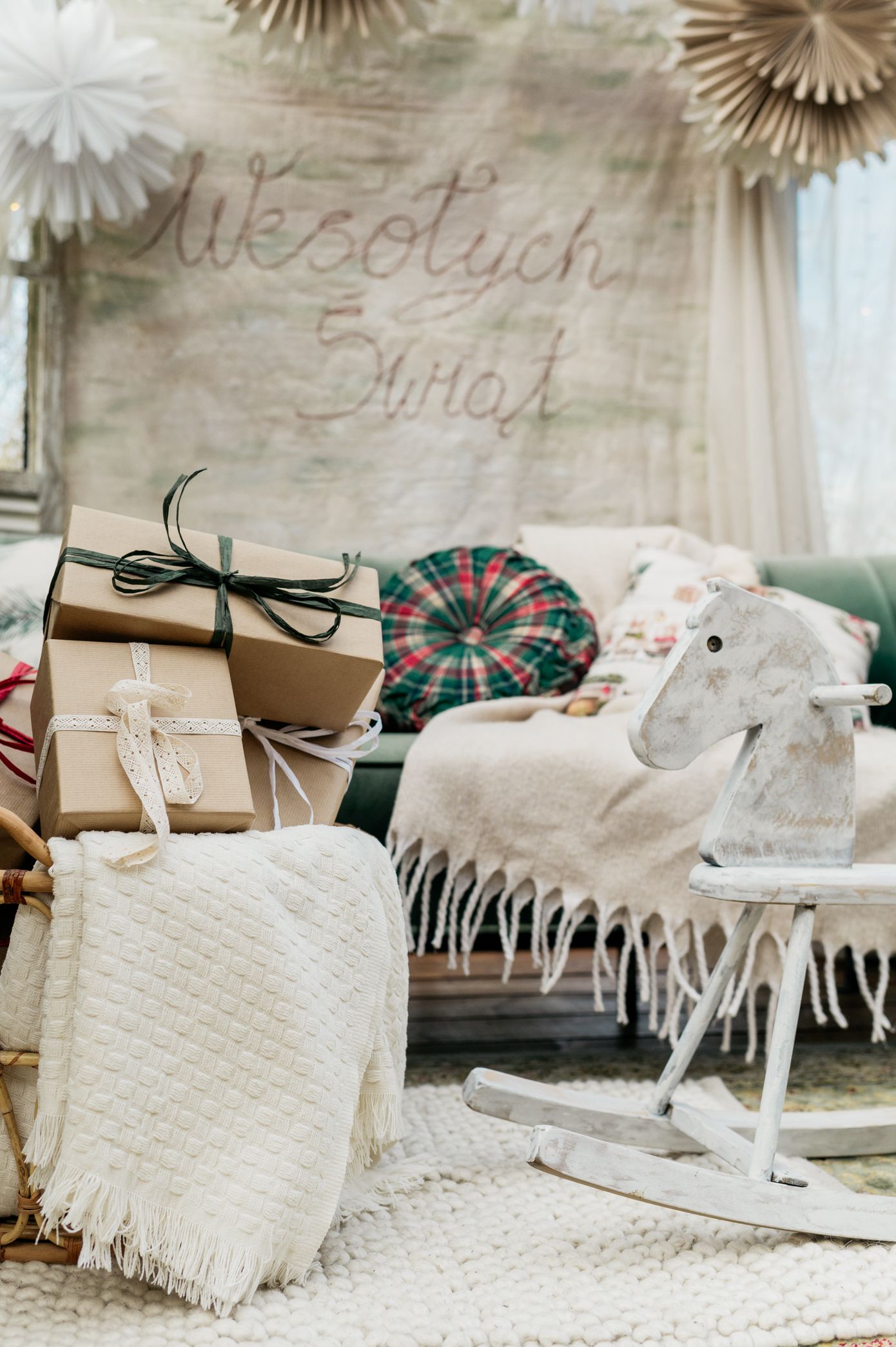 Cozy nursery decor with wrapped gifts, white blankets, plush rocking horse and decorative paper fans on wall.