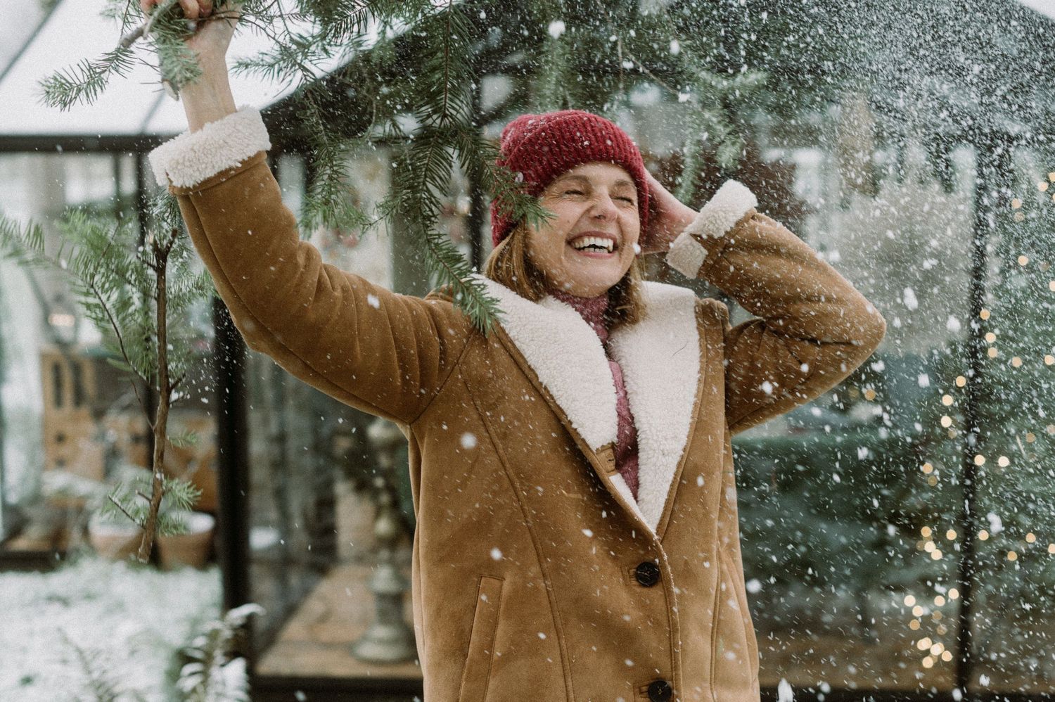 Person in brown coat and red beanie smiles joyfully while playing in falling snow on a winter day.