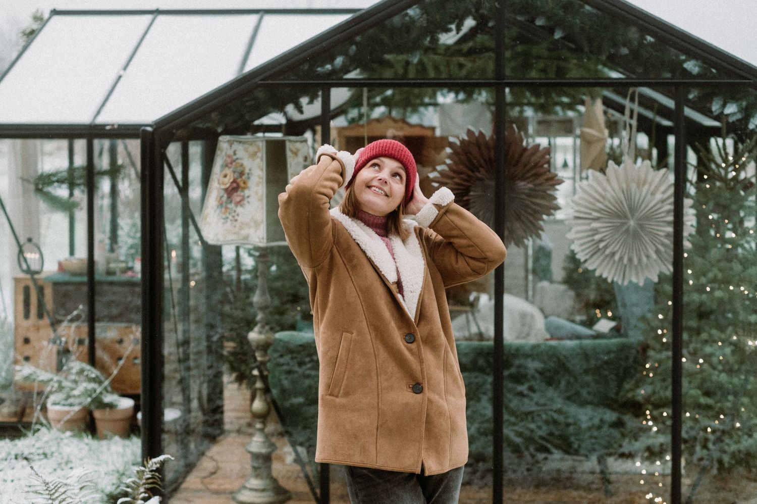 Person in brown coat and red beanie smiles while holding a steaming beverage at a cozy winter garden setup.