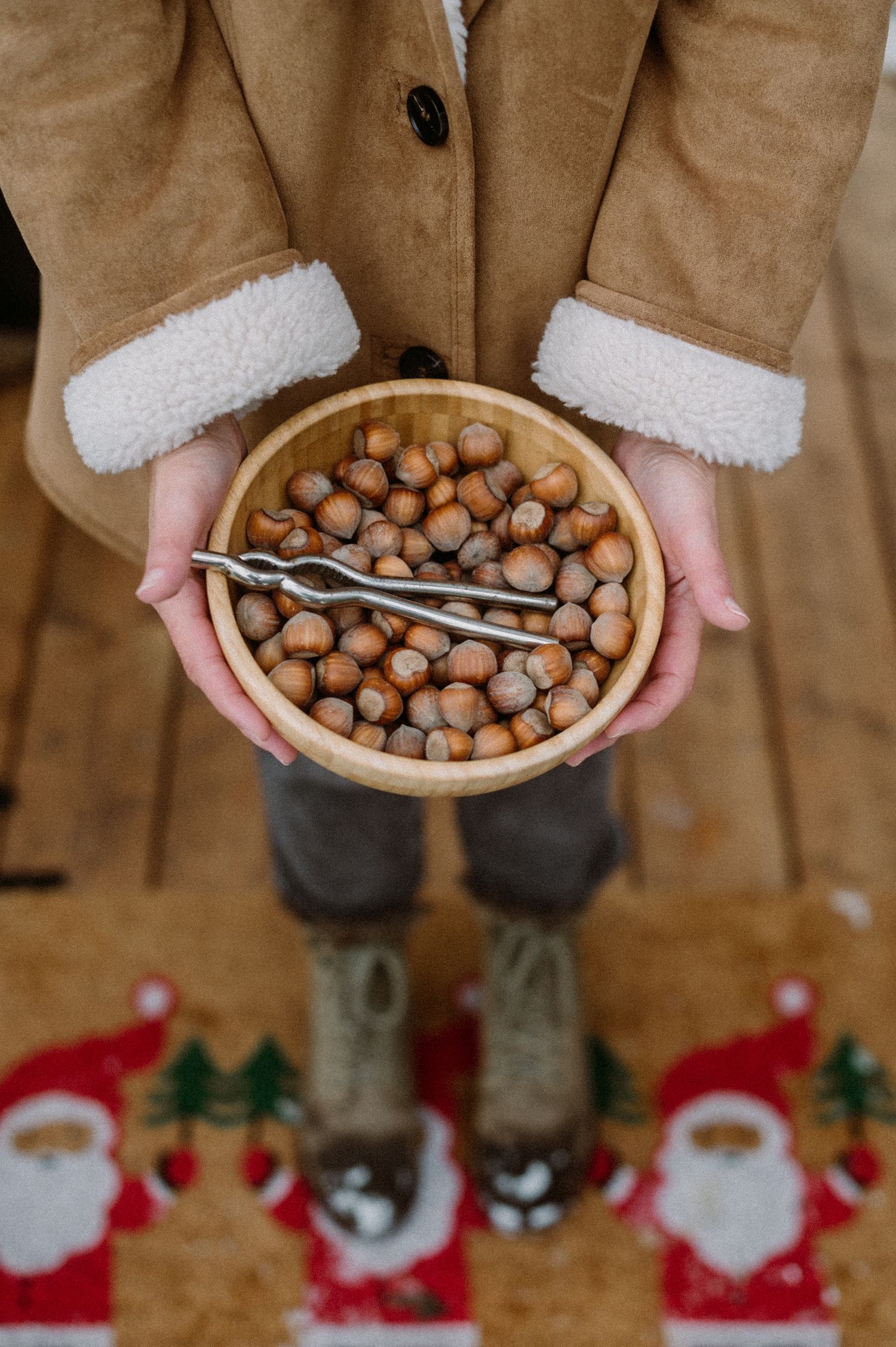 Hands wearing white mittens hold a wooden bowl filled with hazelnuts during Christmas time with holiday decor visible.