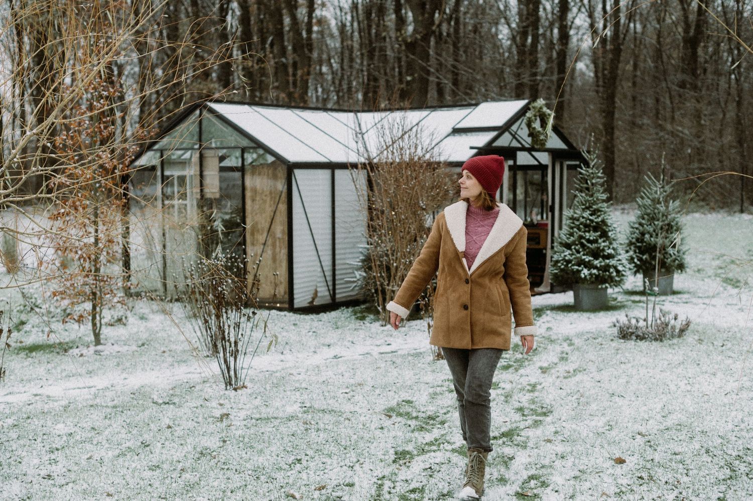 A person in a brown jacket walks through a snowy winter landscape with bare trees and small buildings in the distance.