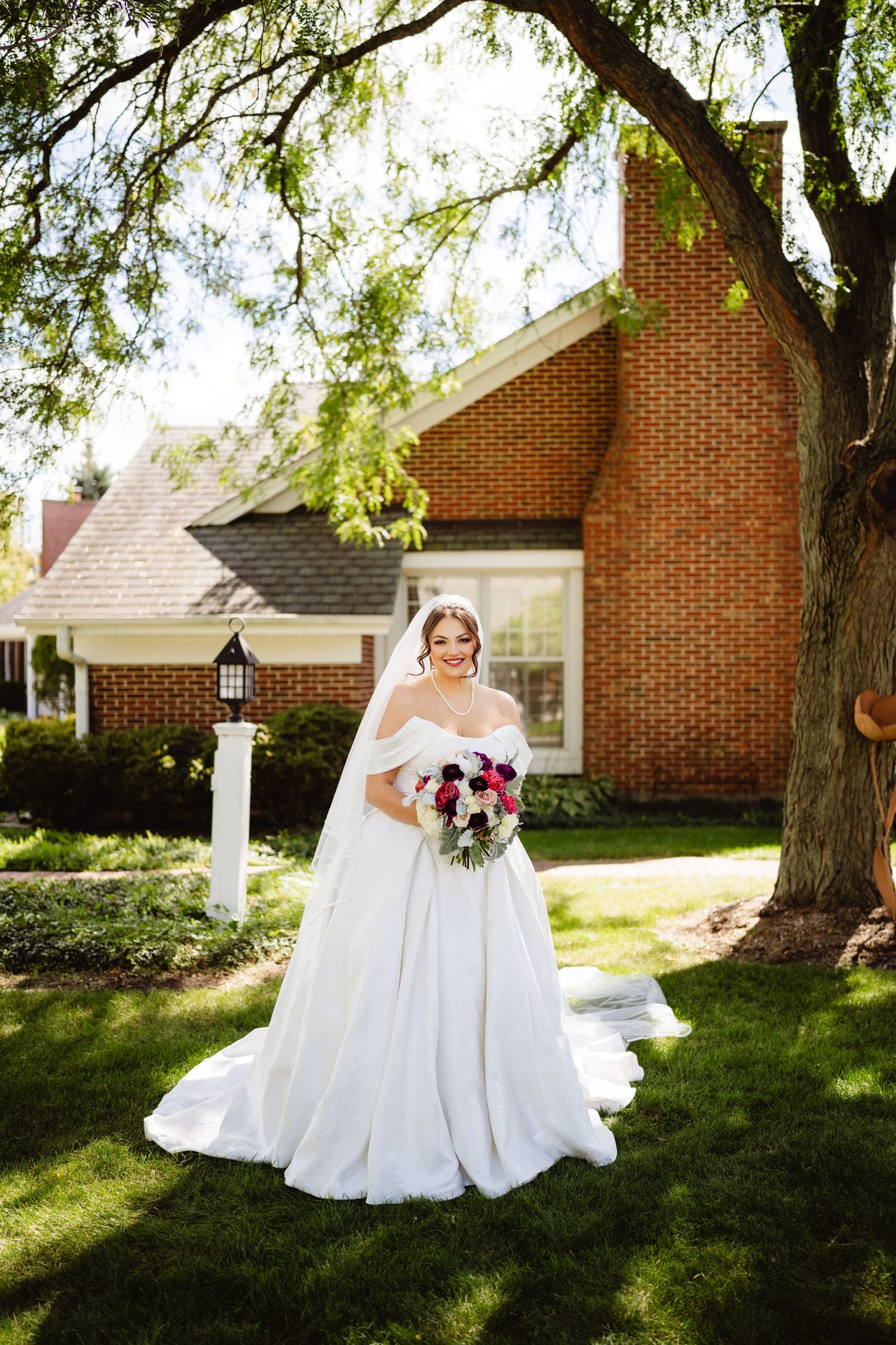 A bride in a flowing white wedding dress sits gracefully on lawn chairs in front of a traditional brick home.