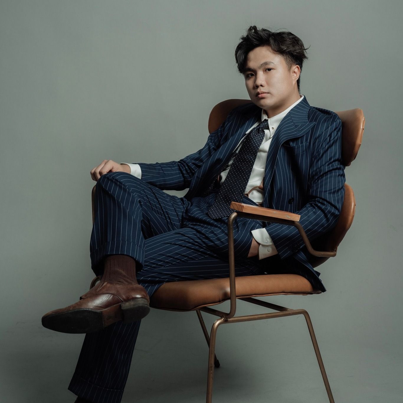 Young man in navy pinstripe suit seated confidently in a leather chair.
