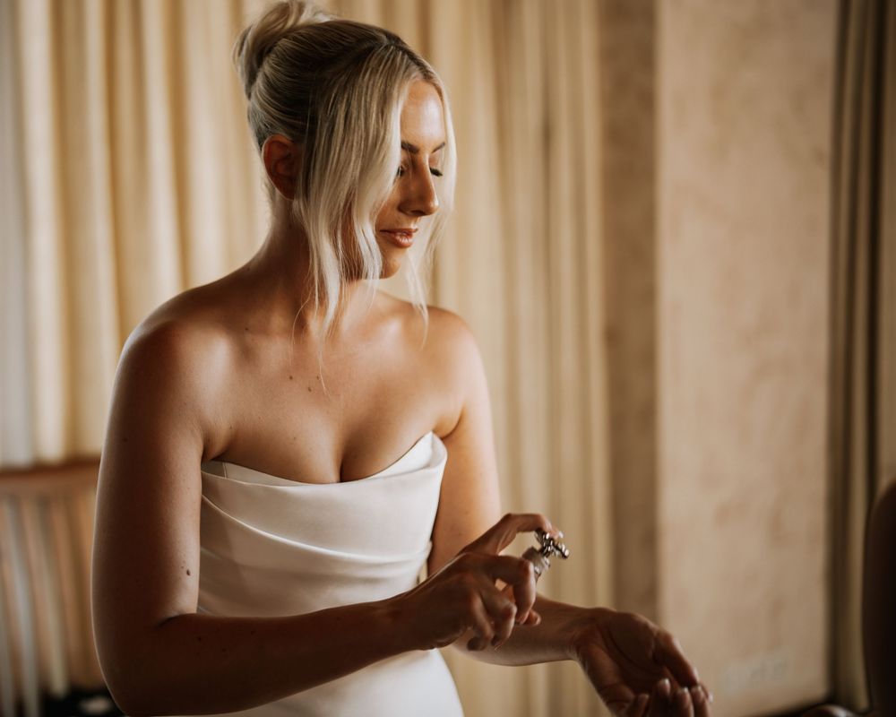 A bride in a white strapless dress stands near a window with soft, warm lighting and neutral-toned curtains in the background.