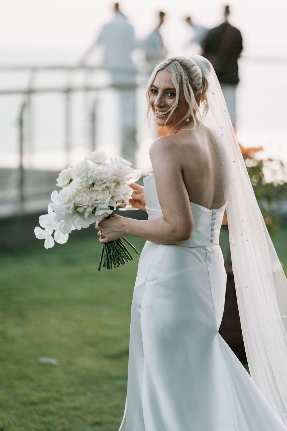 A bride in an elegant white slip dress and flowing veil carries a white orchid bouquet on her wedding day.