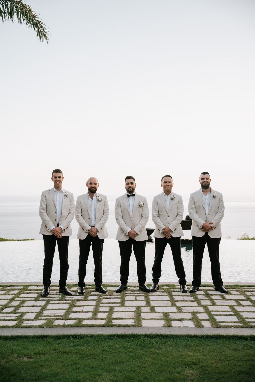 Five groomsmen in matching white dinner jackets stand together on a stone patio overlooking the ocean.