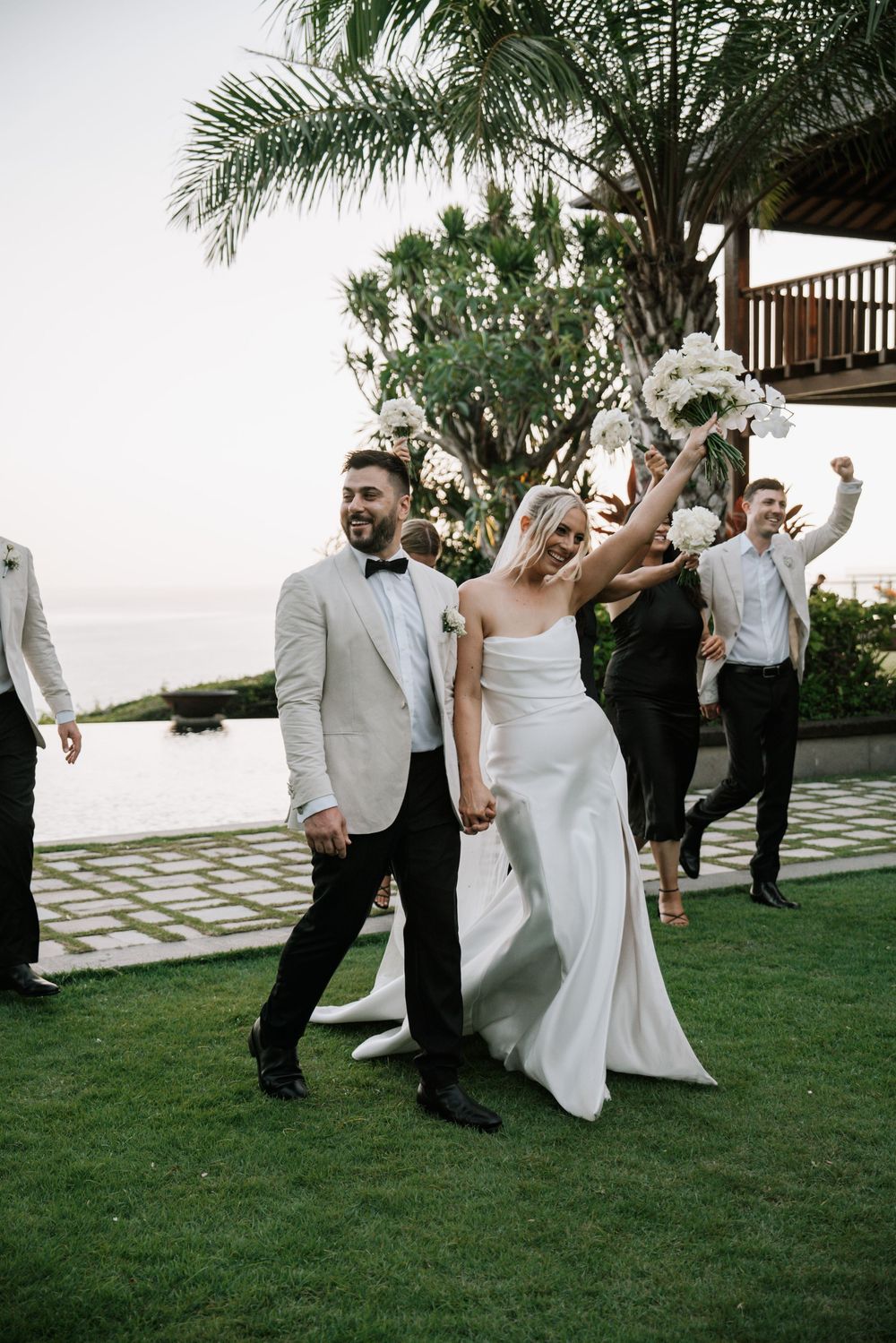 Wedding celebration with guests dancing and cheering on a lawn with palm trees and ocean view in the background.