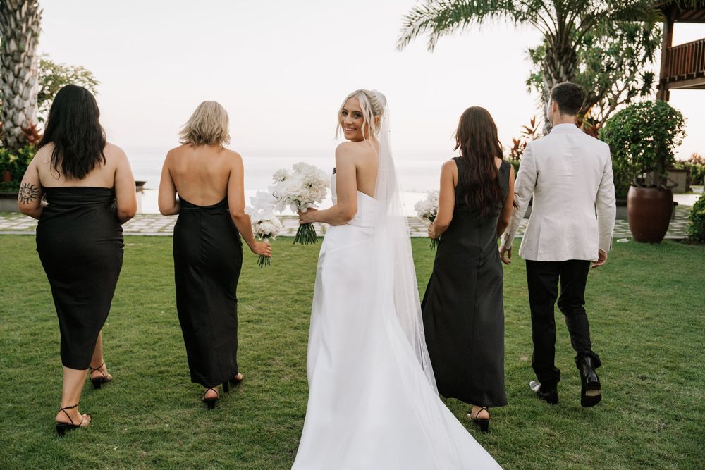 Small group of wedding guests walking single file on a grassy lawn while lined up for photos at outdoor ceremony.