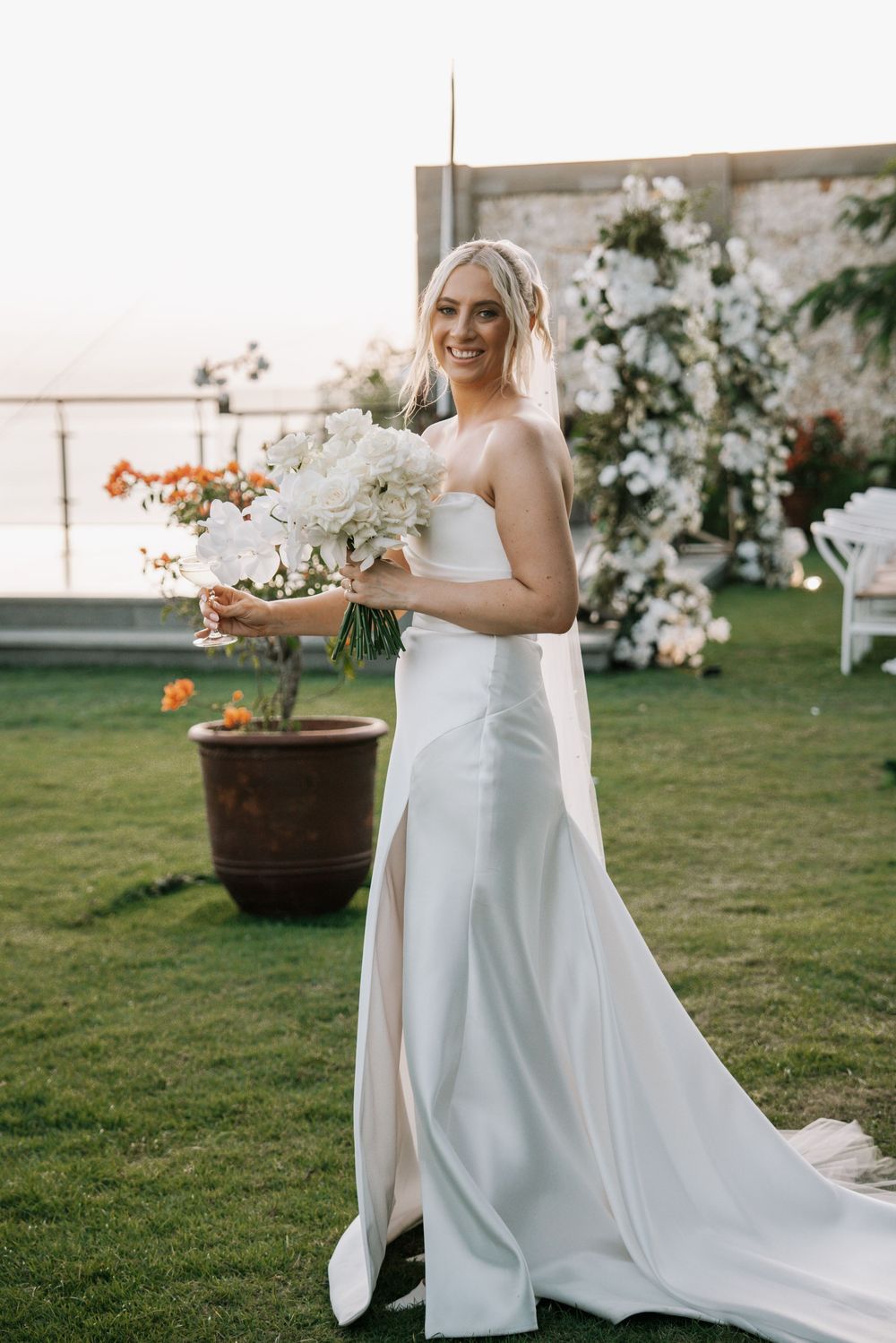 A bride in a flowing white wedding dress holds her bouquet while standing in a grassy outdoor garden setting.