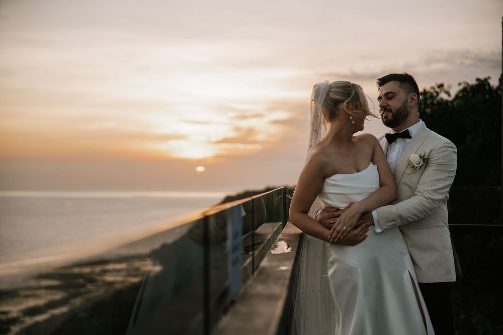 A couple in wedding attire embraces while posing for romantic sunset photos by the ocean.