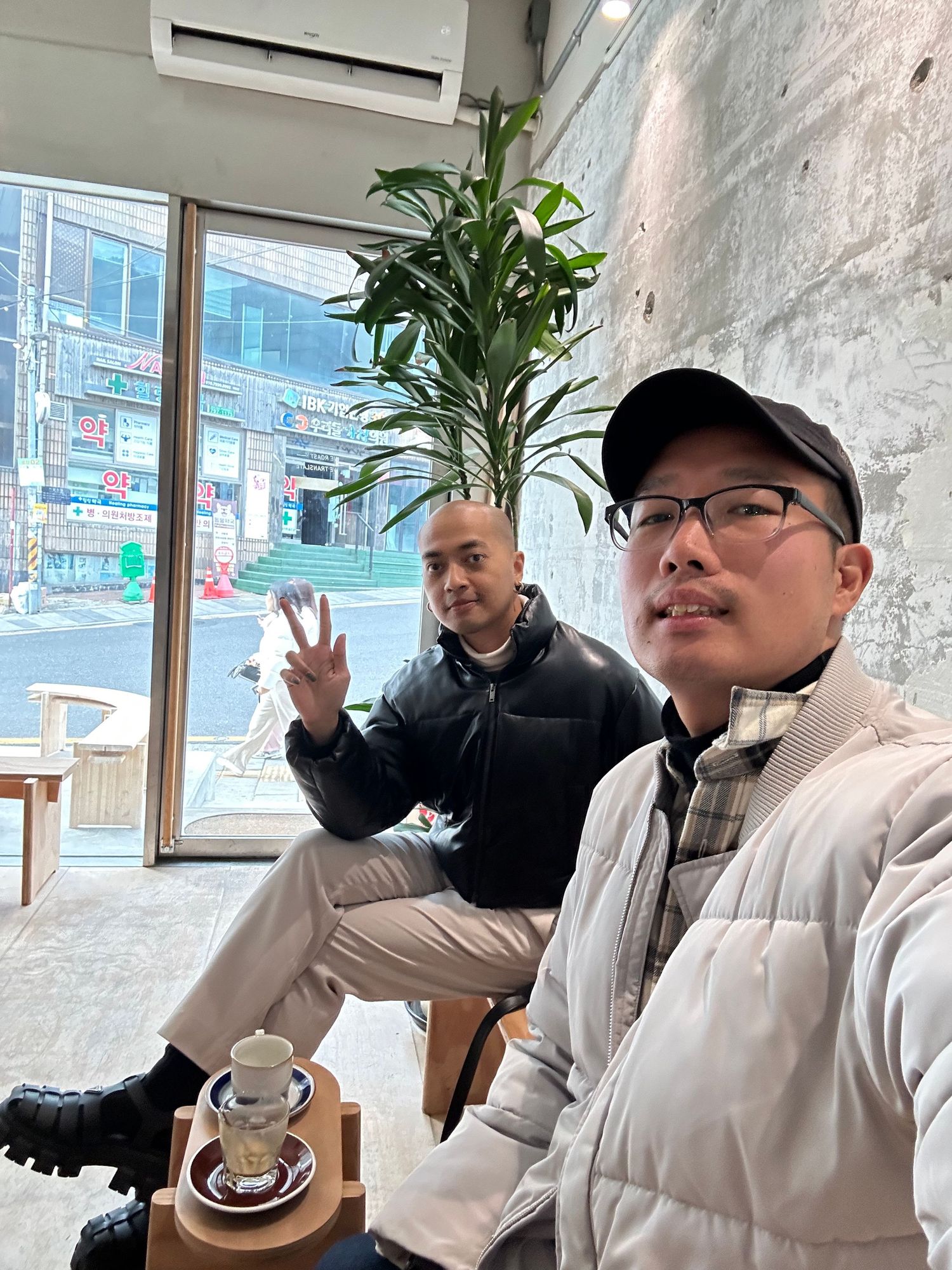 Two people enjoy drinks at a modern cafe with a large window showing an urban street view and a tall indoor plant.