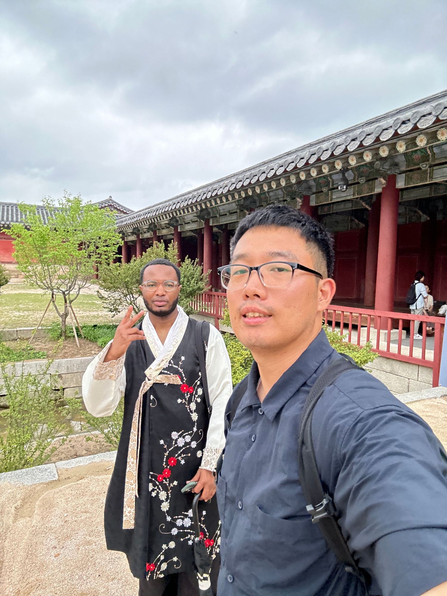 Visitors in traditional hanbok clothing and modern attire explore the historic red-pillared corridors of a Korean palace.