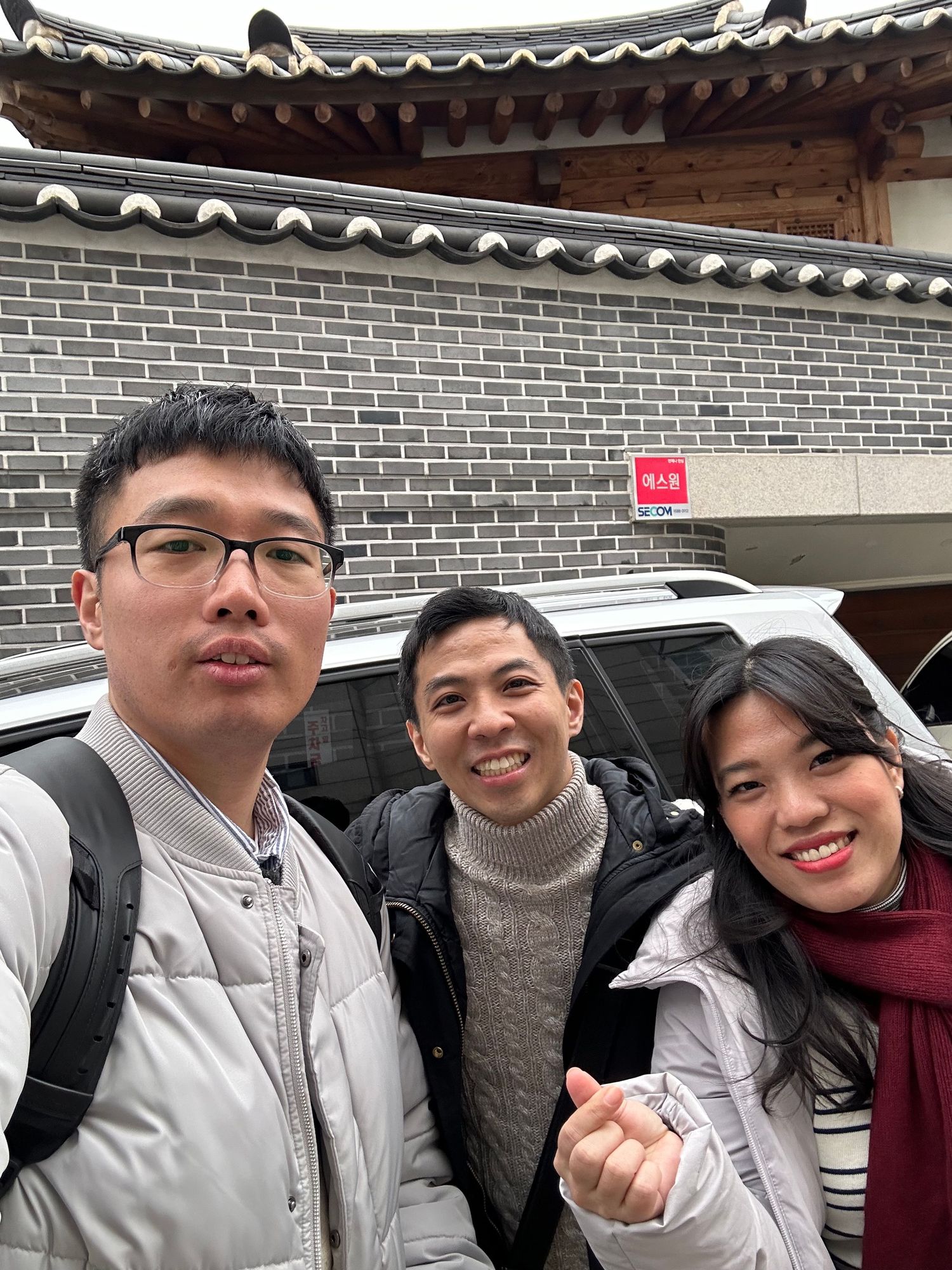 A group of friends in winter clothes smiling together outside a traditional Korean-style building with tiled roof.