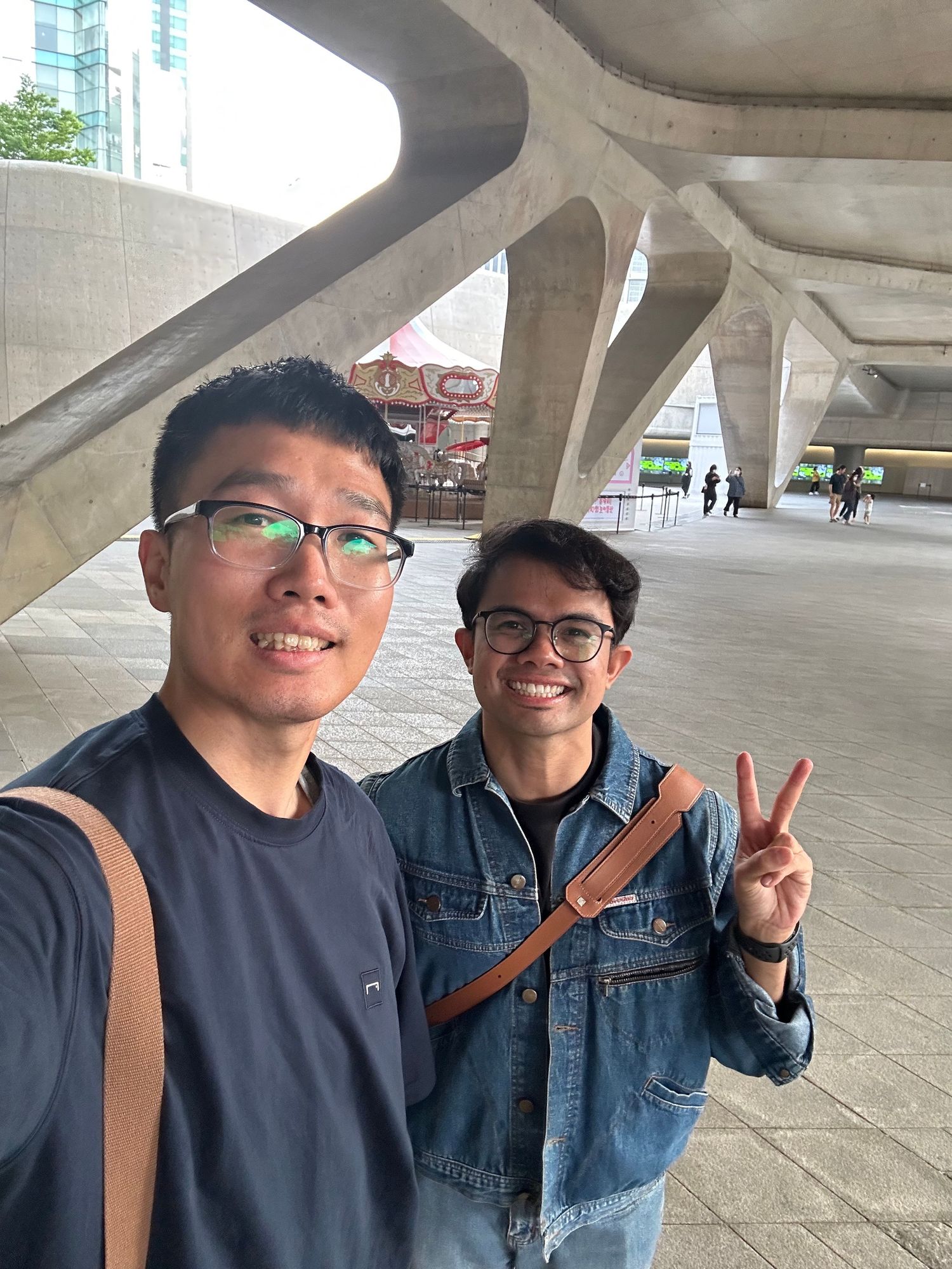 Two friends pose together under a concrete architectural structure making peace signs and showing friendly expressions.