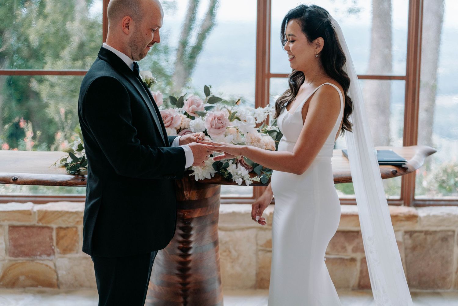 A couple exchanges rings during an intimate wedding ceremony in a sunlit room with stone walls and windows.