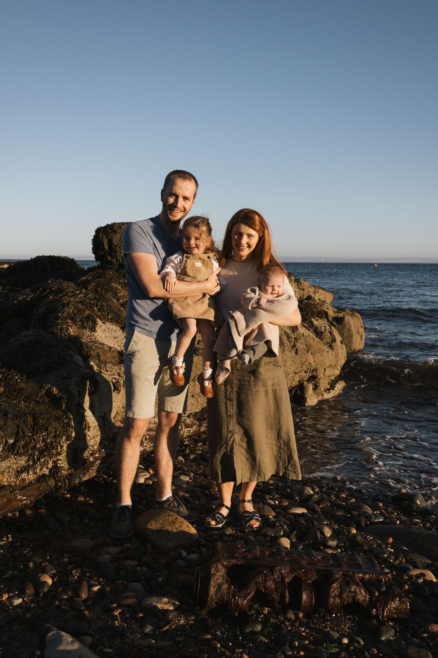 Family sharing a tender moment together on rocky coastal shore during golden hour sunset.