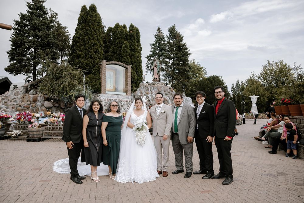 Wedding party poses together on stone patio with evergreen trees and guests in background.