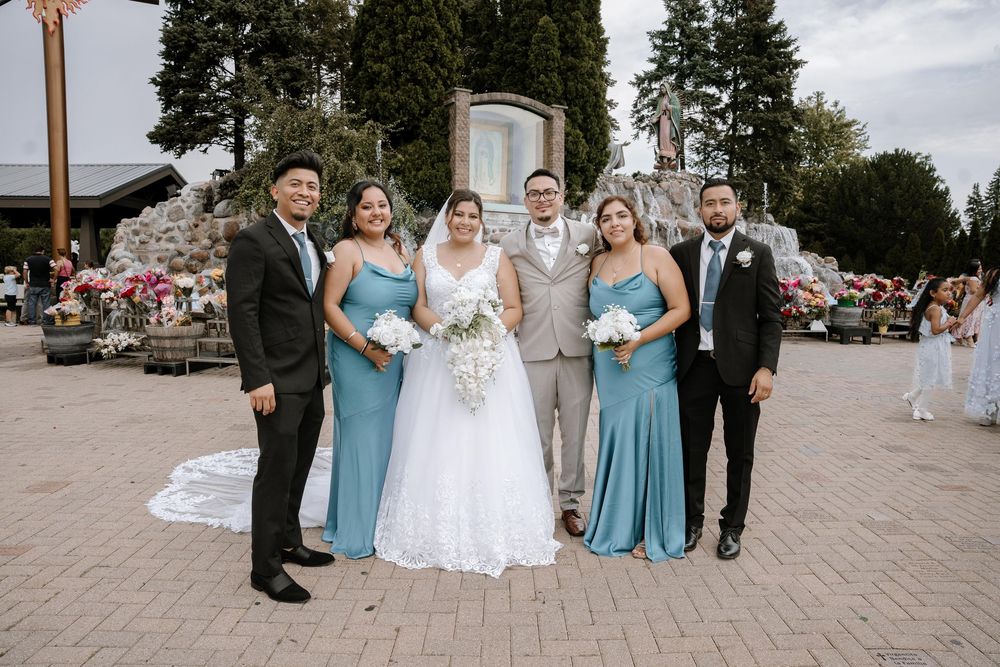 A wedding party poses outdoors in light blue bridesmaid dresses and formal attire against greenery and sky.