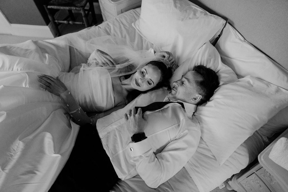 Black and white photo of a just-married couple smiling and cuddling and lying together on a bed in their wedding clothes by August and Vine