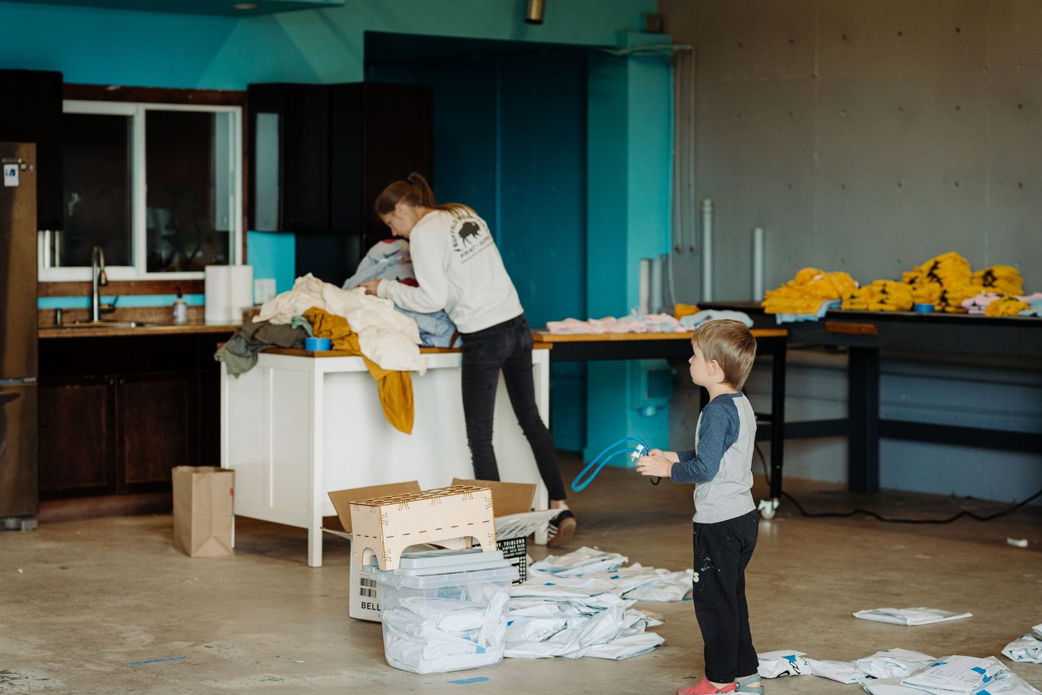 Two people organize and sort through boxes and supplies in a room with turquoise walls and yellow items on shelves.