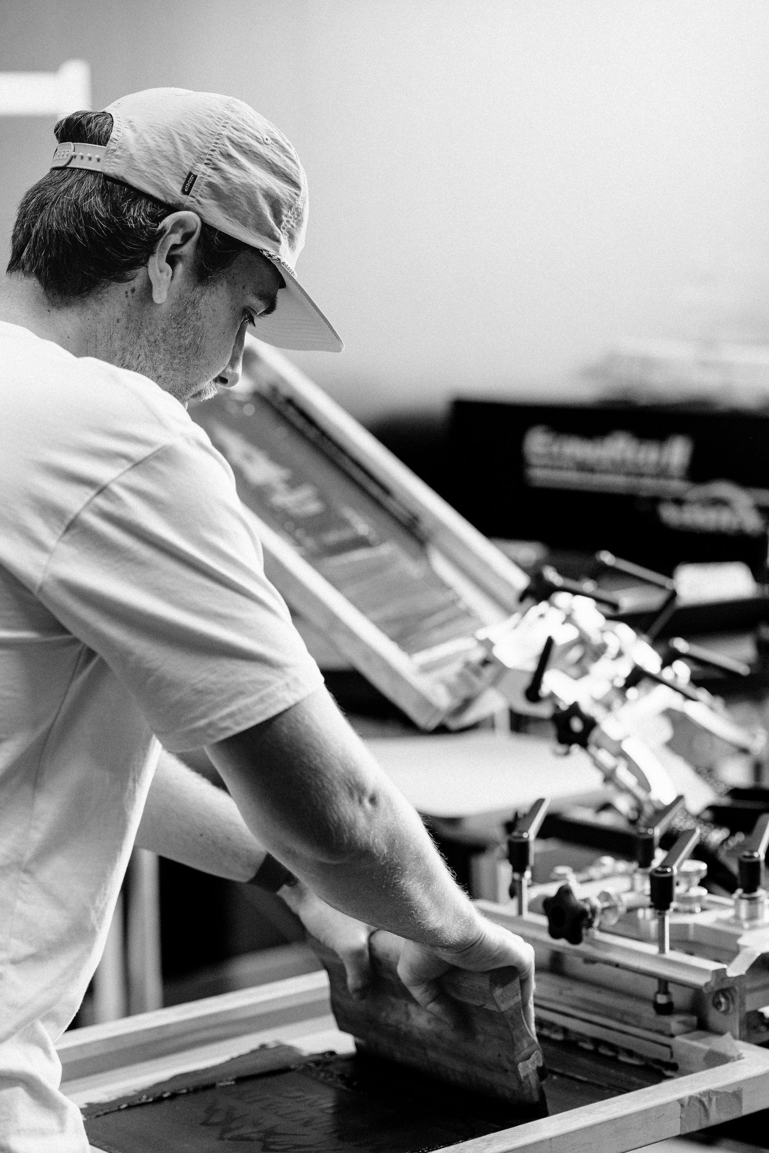Screen printer in a white t-shirt and baseball cap working at a screen printing station using a squeegee tool.