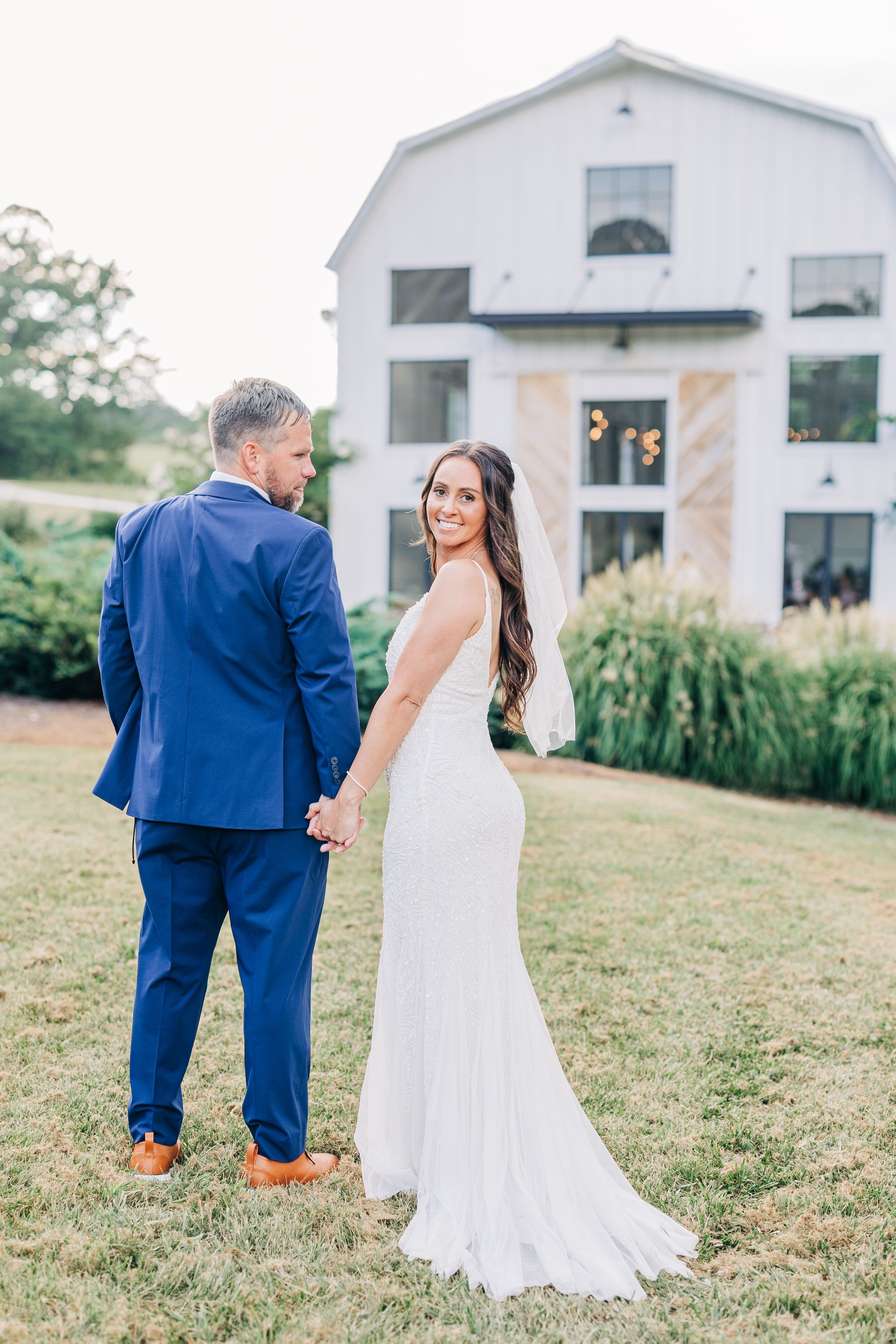 Wedding couple in blue suit and white dress share candid moments while walking on grassy lawn.