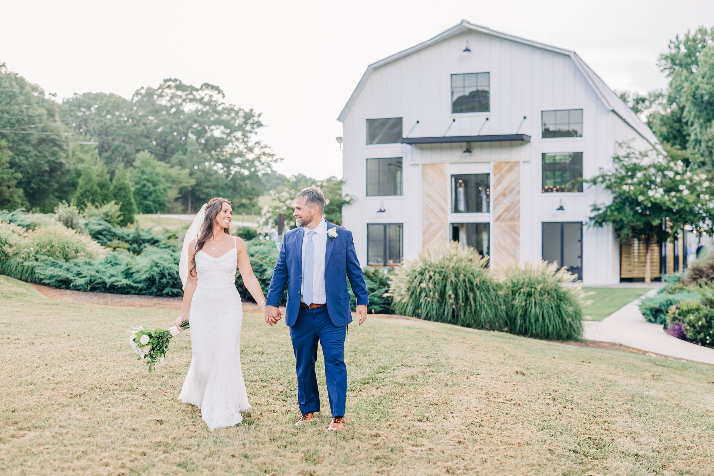 A couple walks hand in hand across a lush green hillside near a white farmhouse on their wedding day, couple portrait at Barn South