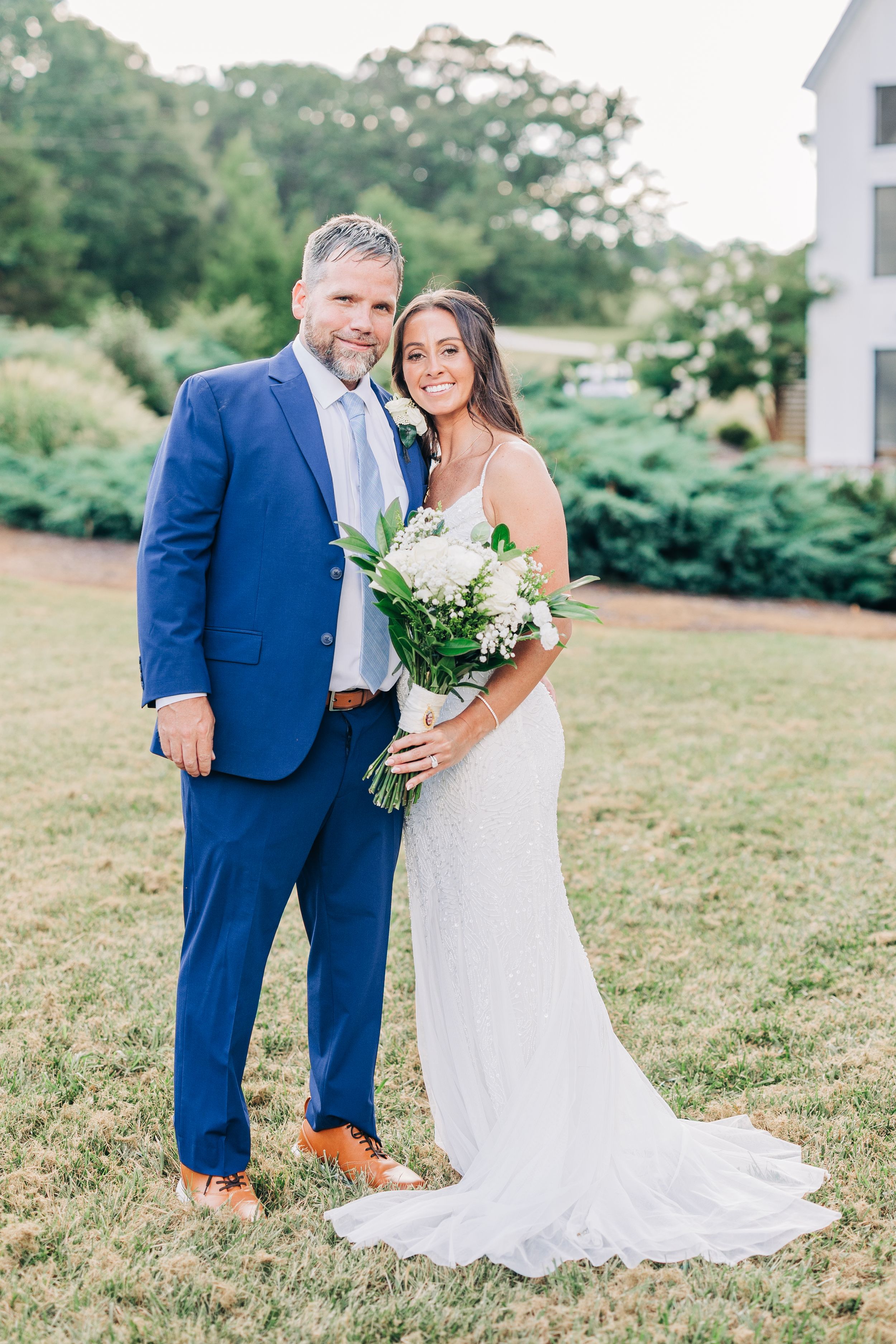 An elegant outdoor wedding photo featuring a couple in a navy suit and white lace gown holding a white floral bouquet.