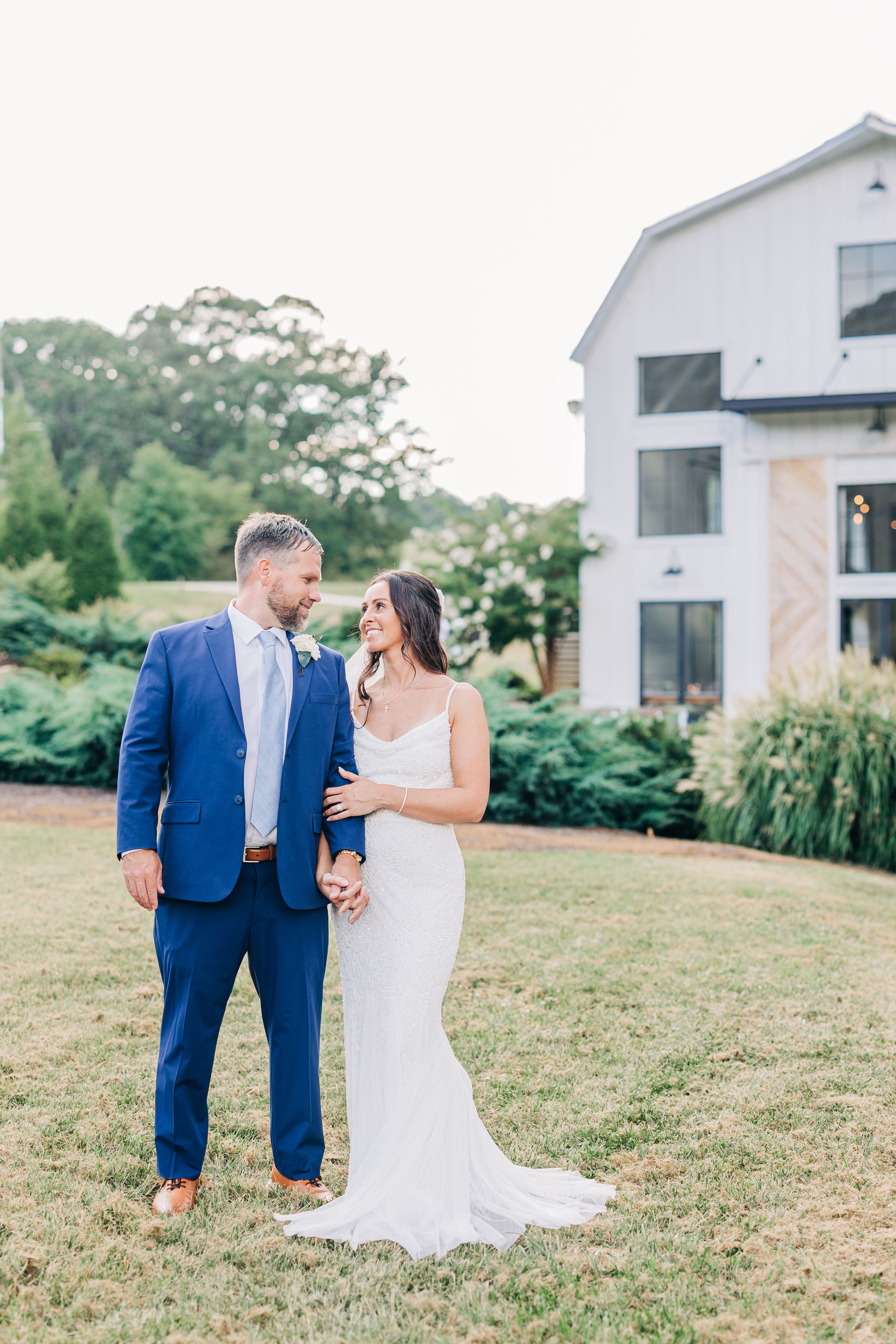 Couple in navy suit and white wedding dress sharing intimate moments on grassy hill near modern white buildings.