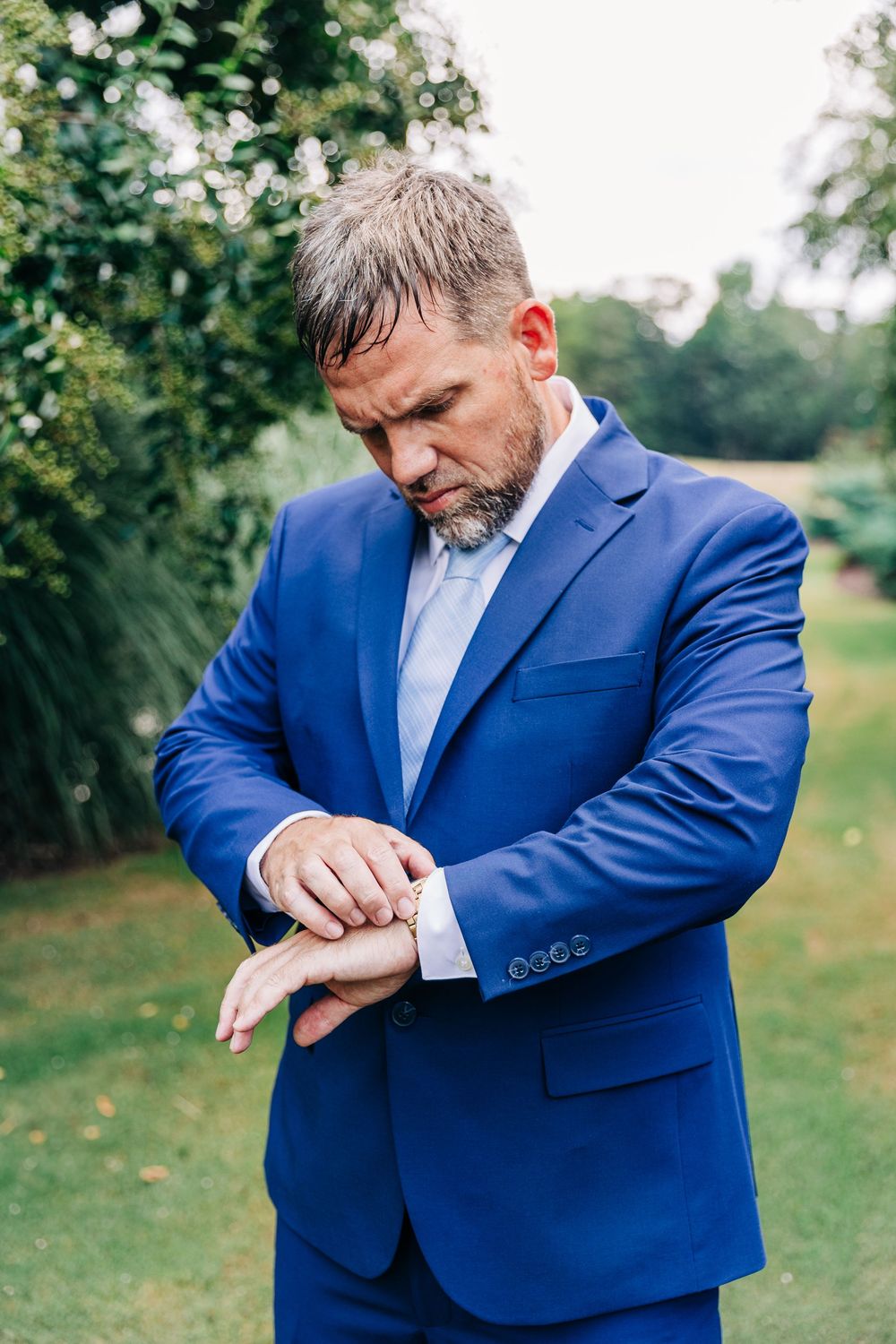 A person in a blue suit checks their wristwatch or smartwatch while standing outdoors in a garden setting.