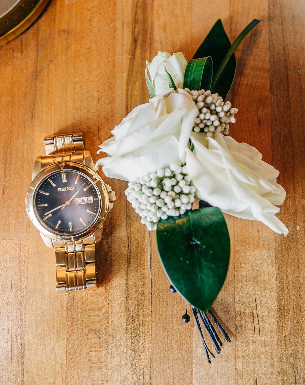 A gold wristwatch and white floral boutonniere with baby's breath and green leaves on a wooden surface.